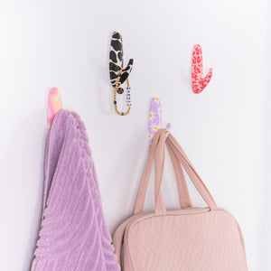 Decorative hooks with unique designs on a white wall, holding a purple garment and beige bag.