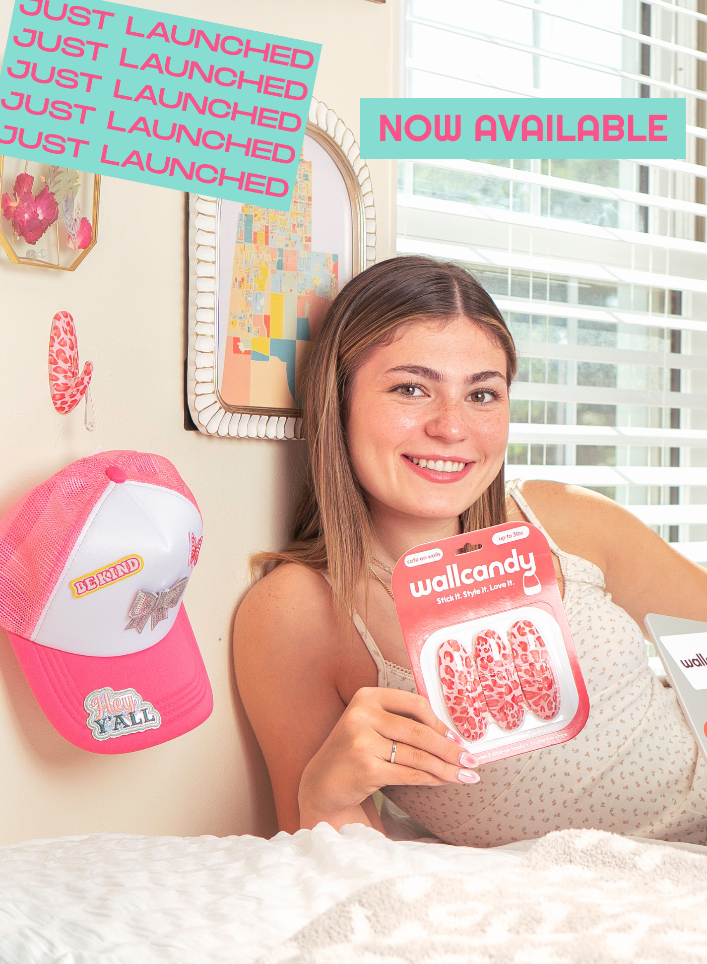 Woman smiling on a bed holding WallCandy Pink Cheetah hooks package, with “Just Launched” and “Now Available” promotional text in the background.
