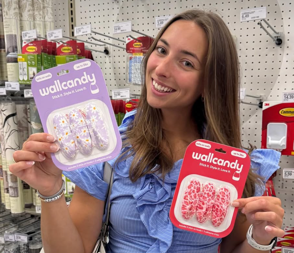 Woman smiling and holding WallCandy adhesive wall hooks in purple flower and pink cheetah designs inside a retail store.