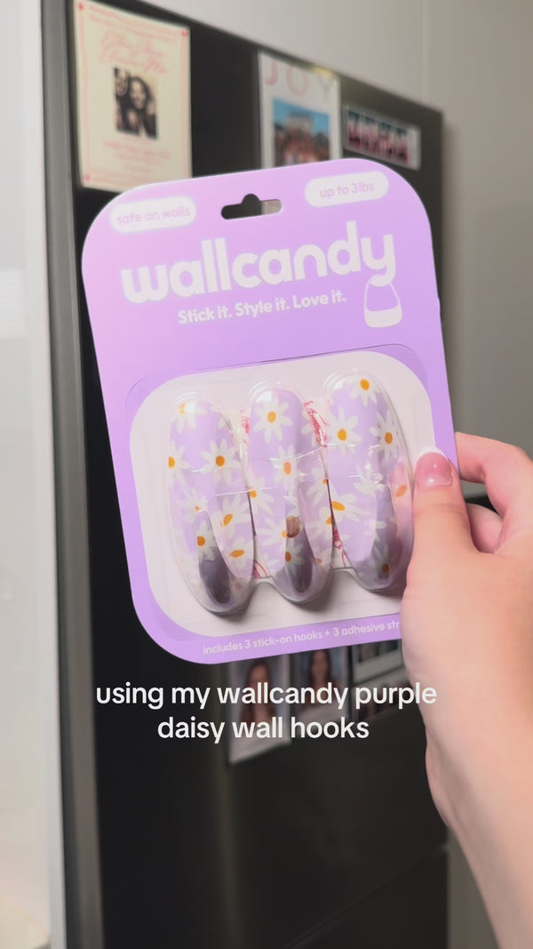 Hand holding a WallCandy Purple Daisy Hook pack (lavender background with white daisy floral design), text overlay says “using my wallcandy purple daisy wall hooks.