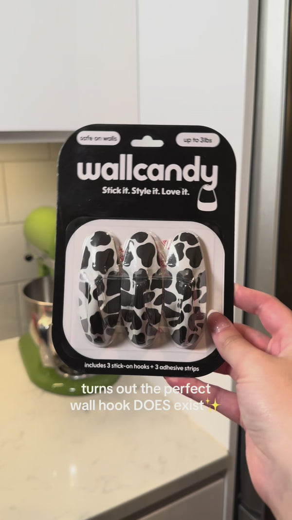 Hand holding a WallCandy Cow Print Hook pack (black and white cow pattern wall hooks with 3 adhesive strips), text overlay says “turns out the perfect wall hook DOES exist ✨