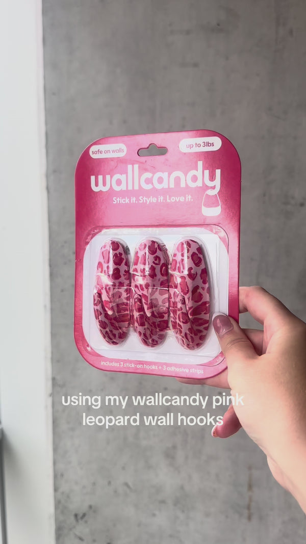 Woman smiling on a bed holding WallCandy Pink Cheetah hooks package, with “Just Launched” and “Now Available” promotional text in the background.